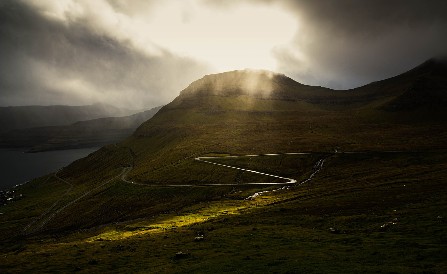 <p>A momentary spotlight of sunshine illuminates the winding road out of Funningur.</p>
